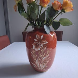 以下が含まれることがあります： 金色と白のフローラルデザインが施された赤い陶器の花瓶。花瓶にはオレンジ色のバラが活けてあります。