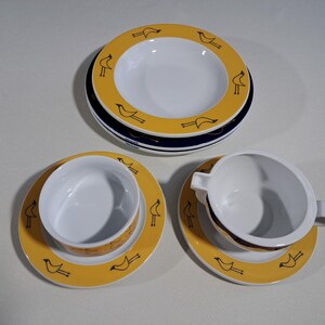 Puede incluir: Un juego de vajilla de cerámica blanca con borde amarillo y siluetas de pájaros negros. El juego incluye platos, cuencos y una taza de sopa con asa. Los platos tienen un diámetro de unos 20 cm.