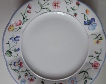 May include: White ceramic plate with a scalloped edge and a light blue border. The plate is decorated with a floral pattern featuring pink, blue, and yellow flowers, along with green leaves and small red berries.