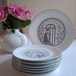 Pode incluir: Um conjunto de seis pratos de cerâmica branca com um desenho cinza e branco de uma mulher com chapéu de pé numa porta. Os pratos estão empilhados uns sobre os outros.