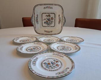 May include: A vintage porcelain serving set featuring a square serving dish with handles and six matching plates. The white dishes have a floral and bird design in the center, with a decorative border. The set is arranged on a white tablecloth.