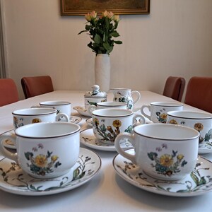 Op de afbeelding: Een set witte theekopjes en schoteltjes met bloemenmotieven. De kopjes en schoteltjes hebben een bruine rand en een bloemenpatroon met groene bladeren en gele bloemen. De set bevat een suikerpot en een roomkan.