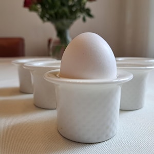 May include: A white ceramic egg cup with a white egg inside. There are three other egg cups in the background.