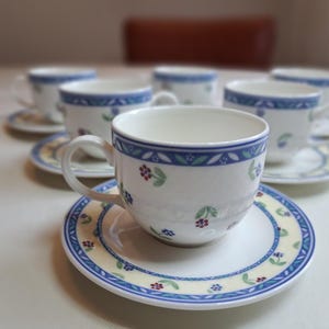 Op de afbeelding: Een set van zes witte keramische theekopjes en schotels. Elk kopje en schotel heeft een blauw en geel bloemmotief rond de rand. De kopjes hebben een gebogen oor en staan op een wit oppervlak.