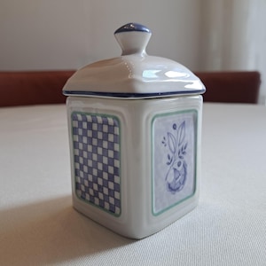 May include: A square ceramic jar with a lid, featuring a white base with blue and green accents. The jar has a checkered pattern on one side and a floral design on another. The lid has a small blue knob.