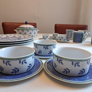 May include: A full porcelain dinner service with a blue and white leaf design. The set includes plates, bowls, cups, saucers, and a lidded serving dish. The rims are accented with a green border.