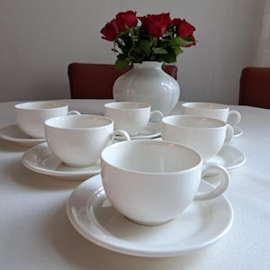 May include: A set of six white ceramic teacups and saucers. The cups are all the same size and shape, and they have a simple, elegant design. The saucers are also white and have a slightly raised edge.