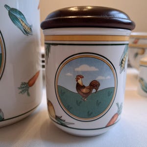May include: A white ceramic canister with a brown lid and a rooster illustration. The canister has a green, yellow, and blue design with a white background. The rooster is red, orange, and yellow.