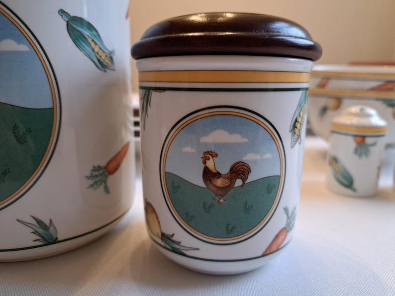 May include: A white ceramic canister with a brown lid and a rooster illustration. The canister has a green, yellow, and blue design with a white background. The rooster is red, orange, and yellow.
