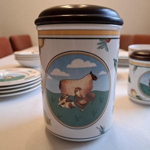 May include: A white ceramic canister with a brown lid, featuring a colorful illustration of a sheep, a rooster, and a rabbit in a green field with blue clouds. The canister has a yellow and gold trim around the top and bottom.