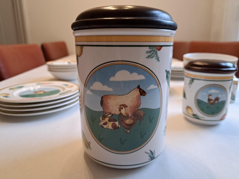 May include: A white ceramic canister with a brown lid, featuring a colorful illustration of a sheep, a rooster, and a rabbit in a green field with blue clouds. The canister has a yellow and gold trim around the top and bottom.