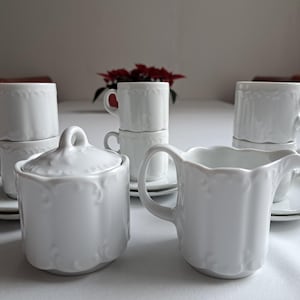 Könnte beinhalten: Ein weißes Porzellan-Tee-Set mit Zuckerdose, Milchkännchen und vier Tassen und Untertassen. Das Set hat ein zartes Blumenmuster.