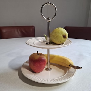 Könnte beinhalten: Eine zweistöckige Etagere aus cremeweißen Keramiktellern mit geripptem Rand und einer zentralen silberfarbenen Metallstange mit einem runden Griff an der Spitze. Auf der oberen Ebene liegt eine grüne Birne, auf der unteren Ebene liegen ein roter Apfel und eine gelbe Banane.