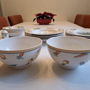 May include: Two white ceramic bowls with a yellow rim and a vegetable design. The bowls are on a white table with other white ceramic dishes.