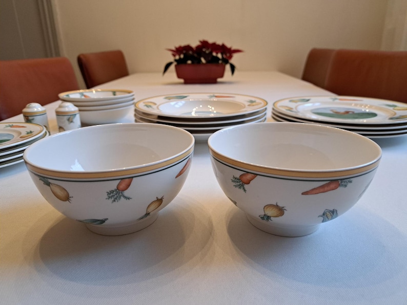May include: Two white ceramic bowls with a yellow rim and a vegetable design. The bowls are on a white table with other white ceramic dishes.