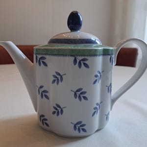 Op de afbeelding: Een witte keramische theepot met een geschulpte vorm en een herhalend patroon van kleine blauwe blaadjes. Het deksel heeft een donkerblauwe knop en een groene band met een gestippelde rand. De theepot is voorzien van een gebogen handvat.