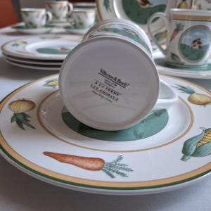 May include: A white ceramic coffee mug with a green and yellow design featuring farm animals and vegetables. The mug has the text "Villeroy & Boch" and "A la Ferme Les Animaux" on the bottom.