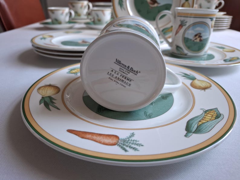 May include: A white ceramic coffee mug with a green and yellow design featuring farm animals and vegetables. The mug has the text "Villeroy & Boch" and "A la Ferme Les Animaux" on the bottom.