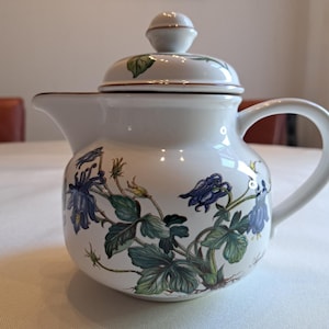 Puede incluir: Tetera de cerámica blanca con tapa y asa. La tetera está decorada con motivos florales azules y verdes. El borde y la tapa están adornados con oro. La tetera está sobre una superficie blanca.