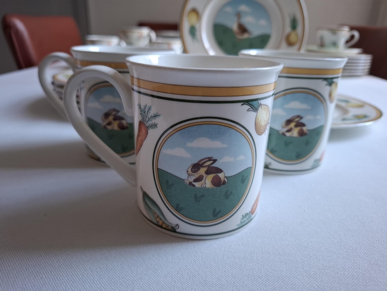 May include: Set of four white ceramic teacups with a green, yellow, and brown floral design featuring rabbits and carrots.