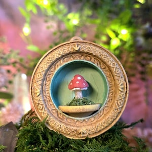May include: A detailed, round, framed art piece featuring a miniature mushroom sculpture. The mushroom has a red cap with white spots and sits on a bed of green moss inside a teal-colored recess. The frame is a light beige with intricate carvings.