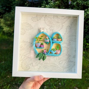 May include: A framed, three-dimensional artwork featuring a light blue shelf design with miniature potted plants and a small white cat figurine. The background has a paisley pattern, and the frame is white.