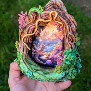 Puede incluir: Escultura de resina de temática fantástica con un paisaje enmarcado con un dragón. El marco está diseñado para parecer un tronco de árbol con follaje verde, flores rosas y una vid amarilla. El paisaje interior representa un dragón cerca del agua.