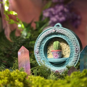 May include: A teal-colored, circular decorative frame with a small cactus in a pot. The frame has an intricate pattern around the edge and a small shelf. A pink and purple crystal is in the foreground, with green moss and foliage.