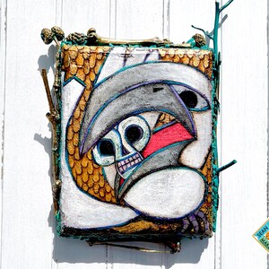 May include: Abstract artwork featuring a skull with a gray hat and a red mouth. The artwork is on a rectangular, textured surface with gold and teal accents. The piece is framed with twigs and has a white background.