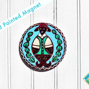 May include: Hand painted magnet featuring a person with a red hat, green shirt, and brown pants. The person is surrounded by green leaves on a blue background. The magnet is round and has a white border.