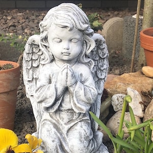 Könnte beinhalten: Eine weiße Betonstatue eines betenden Engels mit Flügeln. Der Engel kniet auf einem Sockel und hat einen ruhigen Gesichtsausdruck.