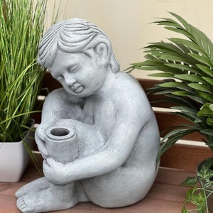 Könnte beinhalten: Eine graue Steinskulptur eines Kindes, das mit gekreuzten Beinen sitzt und ein zylindrisches Objekt auf dem Schoß hält. Die Skulptur hat ein realistisches, detailliertes Gesicht und ist eine dekorative Gartenstatue.