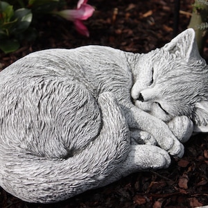 Könnte beinhalten: Eine graue Betonkatzenstatue, die sich in Schlafposition zusammengerollt hat. Die Statue ist mit Fellstruktur detailliert und hat ein realistisches Aussehen.