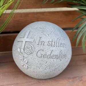 Può includere: Una sfera di pietra grigia con una croce, una rosa e il testo "In stillem Gedenken" inciso su di essa.