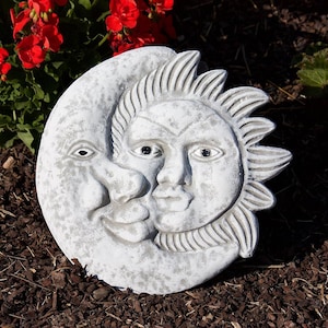 Pode incluir: Um ornamento de jardim de pedra cinza desgastada com um design de sol e lua sorrindo. O sol tem um contorno detalhado com raios, e a lua tem um rosto sorridente. O ornamento está sobre cobertura morta marrom escura, com flores vermelhas ao fundo.