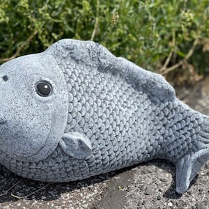 Könnte beinhalten: Eine graue Betonskulptur eines Fisches mit detaillierten Schuppen und einem realistischen Ausdruck. Der Fisch liegt auf der Seite mit leicht geöffnetem Maul.