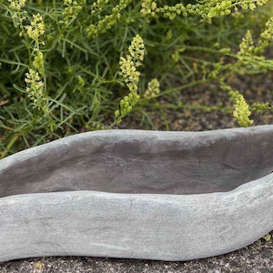 Op de afbeelding: Een grijze, langwerpige plantenbak met een getextureerd oppervlak. De binnenkant is donkerder grijs. Groen gebladerte met kleine gele bloemen is op de achtergrond. Deze plantenbak is geschikt voor diverse planten en bloemen.