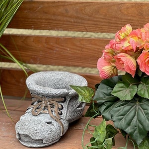Könnte beinhalten: Ein grauer, steinähnlicher Blumentopf in Form eines Stiefels mit braunen Schnürsenkeln. Der Topf ist mit Erde gefüllt und steht neben einem Topf mit rosa und gelben Blumen und grünem Laub. Der Topf steht auf einer Holzoberfläche.