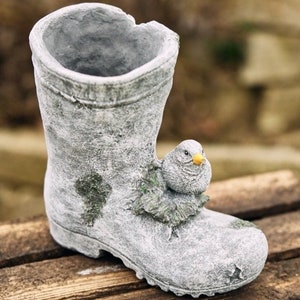 Op de afbeelding: Een grijze betonnen tuinlaars plantenbak met een kleine vogel die op de zijkant zit. De laars heeft een verweerde afwerking en een verweerde uitstraling.