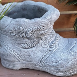 Op de afbeelding: Een grijze betonnen laars plantenbak met een gedetailleerd ontwerp van een slang en andere decoratieve elementen. De plantenbak heeft een brede opening aan de bovenkant om te planten.