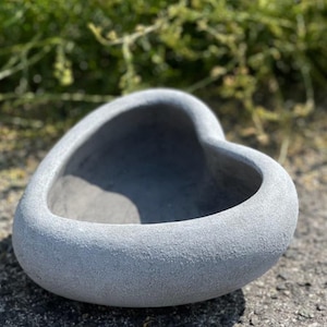 Könnte beinhalten: Ein grauer herzförmiger Pflanztopf aus Beton. Der Topf hat eine raue, strukturierte Oberfläche und einen glatten, abgerundeten Innenraum.