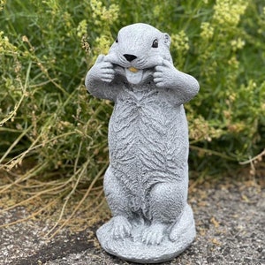 May include: A gray, stone-like statue of a groundhog, with its paws on its cheeks, pulling its mouth wide. The groundhog has a small, yellow tongue visible. The statue is standing on a stone base, with a textured surface.