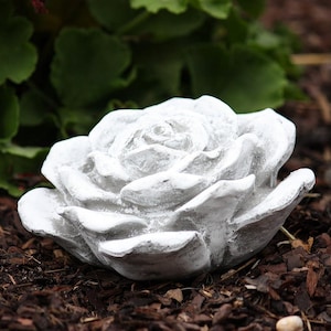 Könnte beinhalten: Eine detaillierte, weiße Rosen-Skulptur aus Stein. Die Blüte ist voll erblüht, mit mehreren Schichten von Blütenblättern. Die Skulptur steht auf einer dunklen, braunen Mulchschicht, mit grünem Laub im Hintergrund.