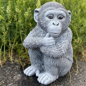 Könnte beinhalten: Eine graue Betonstatue eines Affen, der mit gekreuzten Händen sitzt. Der Affe hat eine realistische Fellstruktur und große, ausdrucksstarke Augen.