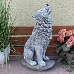 Könnte beinhalten: Eine graue Wolfsstatue aus Stein in heulender Pose. Die Skulptur weist detailliertes Fell auf und steht auf einem Steinsockel. Der Wolf ist vor einer Holzbank und Topfpflanzen positioniert.
