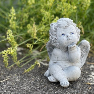 Könnte beinhalten: Eine kleine, graue Steinengelfigur mit Flügeln, die mit dem Kopf nach oben geneigt und einer Hand am Gesicht sitzt. Der Engel hat blaue Augen und einen süßen Ausdruck.