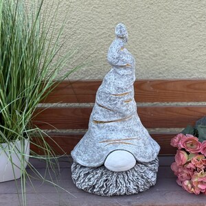 Könnte beinhalten: Eine graue Gartenzwerg-Statue mit einem langen, spiralförmigen Hut und einem weißen Bart. Der Hut hat goldene Akzente. Der Zwerg steht neben einer Topfpflanze und einem Strauß rosa Blumen.