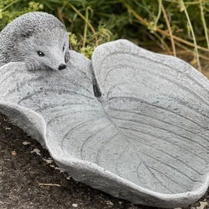 Può includere: Un abbeveratoio per uccelli in cemento grigio a forma di foglia con una piccola statuetta di riccio in cima.
