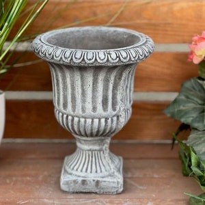 Op de afbeelding: Een grijze betonnen plantenbak met een geribbeld ontwerp en een decoratieve rand. De plantenbak staat op een houten oppervlak.