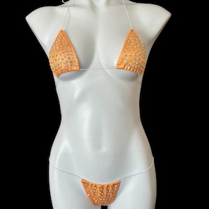 Könnte beinhalten: Ein zweiteiliger Badeanzug in leuchtend orange. Ober- und Unterteil sind dreieckig und haben transparente Träger. Der Stoff ist mit kleinen, schimmernden Strasssteinen besetzt.